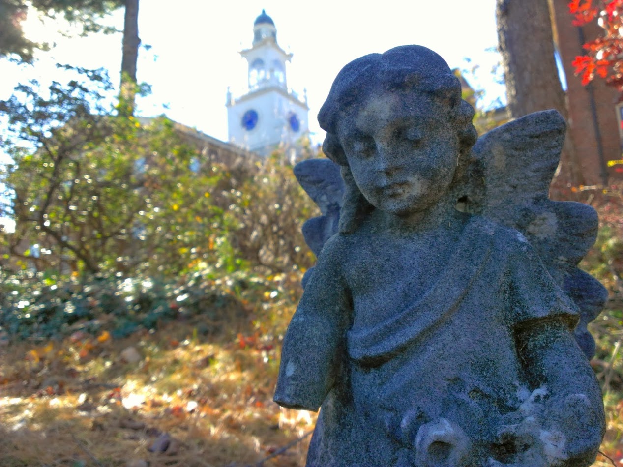 A youthful angel sculpted in stone stands in the foreground; in the background, autumnal trees and the spire of Samuel Phillips Hall can be seen.