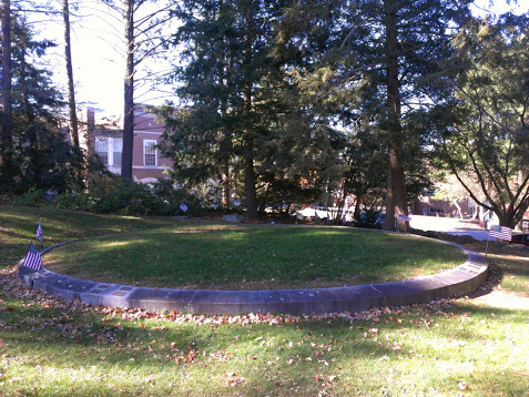 A photograph of a large, circular mound inlaid with bricks around its perimeter and elevated about a foot above the rest of the terrain. Small American flags are planted around the circumference of the garth and pine trees occlude George Washington Hall in the background.