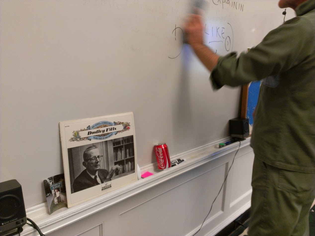 A photograph depicting a vinyl record sleeve bearing the title 'Dudley Fitts' and a picture of the man. The sleeve is resting on the mantle of a whiteboard next to various objects, and a person in a green shirt and pants is erasing some illegible writing on the board.