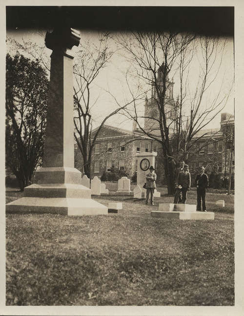 A black-and-white photograph dated from 1928 depicting the 12-foot-tall granite cross memorializing Harriet Beecher Stowe, with three men dressed in suits standing diminuitively in the distance, while several barren trees and Samuel Phillips Hall lie in the background.