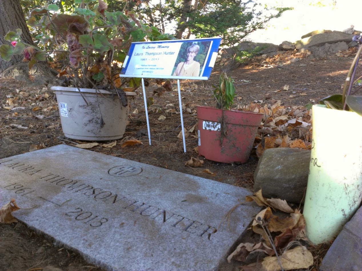A low-angle photograph of a square gravestone laid into the ground, bearing the name Laura Thompson Hunter. Above the stone is an informational sign with a photograph and some biographical details, while a variety of potted plants rest beside it.