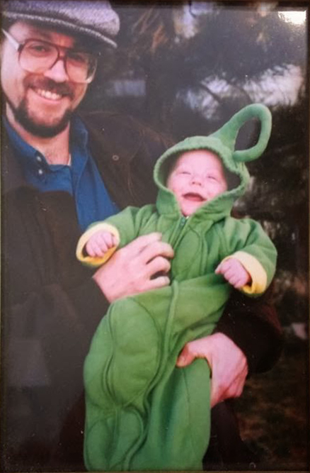 A picture of a framed photograph showing a bearded, bespectacled man holding his very young baby, who is wearing a pea pod Halloween costume. They are both smiling widely.